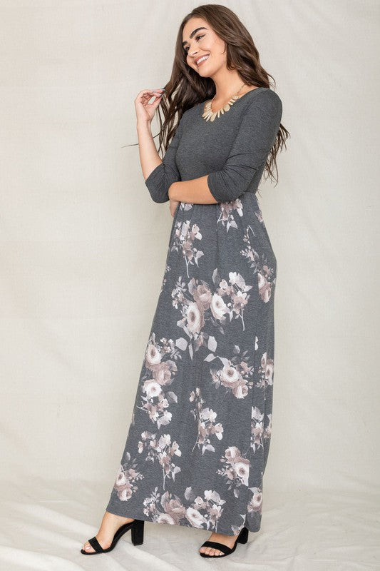 A woman modeling the Quarter Sleeve Floral Maxi Dress, featuring a grey upper section and a floral skirt, stands with one hand on her hip against a plain white background.