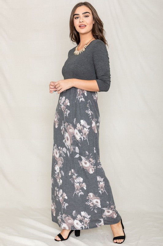 A woman modeling the Quarter Sleeve Floral Maxi Dress, featuring a grey upper section and a floral skirt, stands with one hand on her hip against a plain white background.