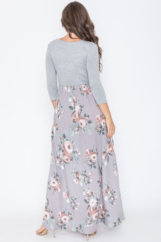 A woman modeling the Quarter Sleeve Floral Maxi Dress, featuring a grey upper section and a floral skirt, stands with one hand on her hip against a plain white background.