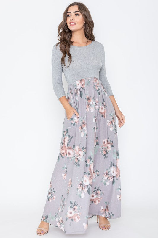 A woman modeling the Quarter Sleeve Floral Maxi Dress, featuring a grey upper section and a floral skirt, stands with one hand on her hip against a plain white background.