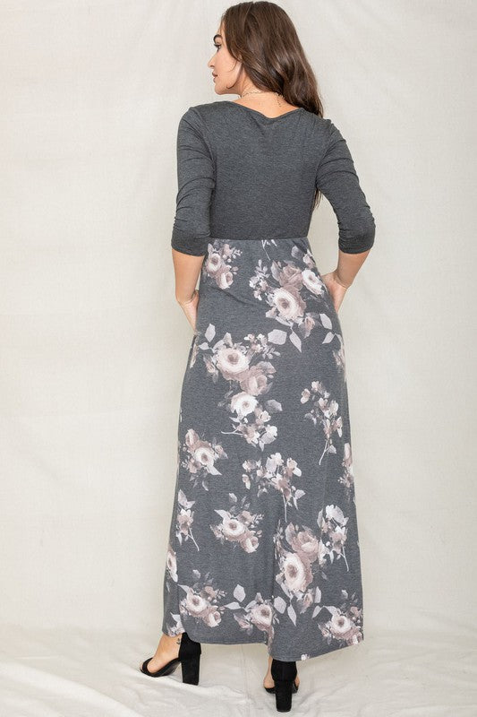 A woman modeling the Quarter Sleeve Floral Maxi Dress, featuring a grey upper section and a floral skirt, stands with one hand on her hip against a plain white background.