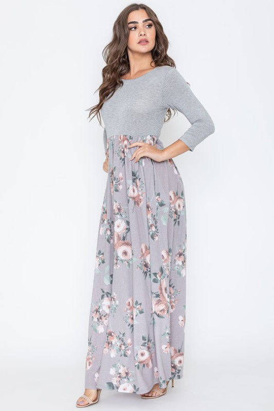 A woman modeling the Quarter Sleeve Floral Maxi Dress, featuring a grey upper section and a floral skirt, stands with one hand on her hip against a plain white background.