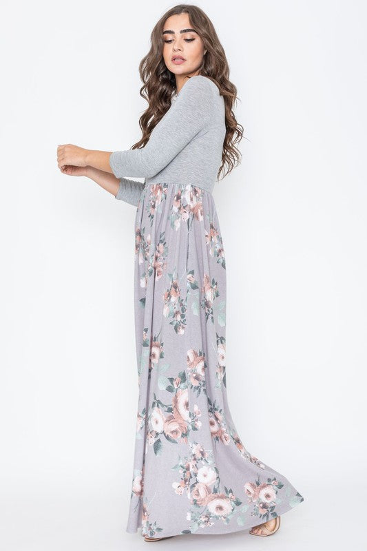 A woman modeling the Quarter Sleeve Floral Maxi Dress, featuring a grey upper section and a floral skirt, stands with one hand on her hip against a plain white background.