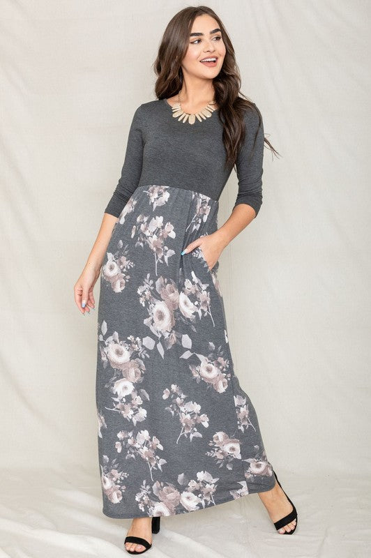 A woman modeling the Quarter Sleeve Floral Maxi Dress, featuring a grey upper section and a floral skirt, stands with one hand on her hip against a plain white background.