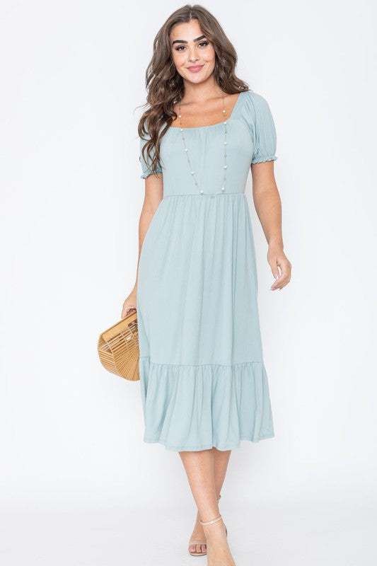 A woman in a Square Neck Puff Sleeve Boho Dress in light blue holds a wooden handbag. She stands on a plain background, wearing strappy heels.