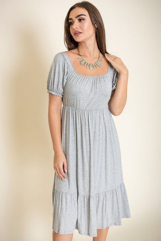 A woman in a Square Neck Puff Sleeve Boho Dress in light blue holds a wooden handbag. She stands on a plain background, wearing strappy heels.