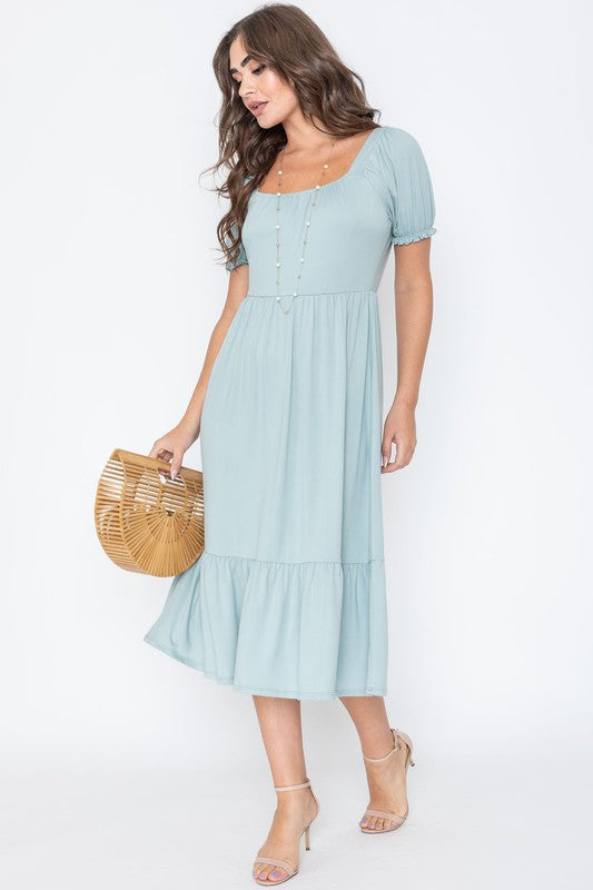 A woman in a Square Neck Puff Sleeve Boho Dress in light blue holds a wooden handbag. She stands on a plain background, wearing strappy heels.