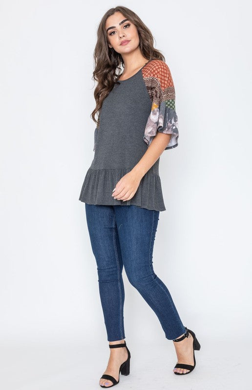 A woman wearing the Patchwork Ruffle Sleeve Tunic, featuring colorful patchwork sleeves, paired with blue jeans, stands against a plain background.
