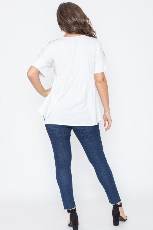 A woman stands against a white background, dressed in the vibrant Tropical Keyhole Tie Front Tunic adorned with a colorful leaf pattern, blue jeans, and black heels. Made in the United States, her ensemble radiates effortless charm.