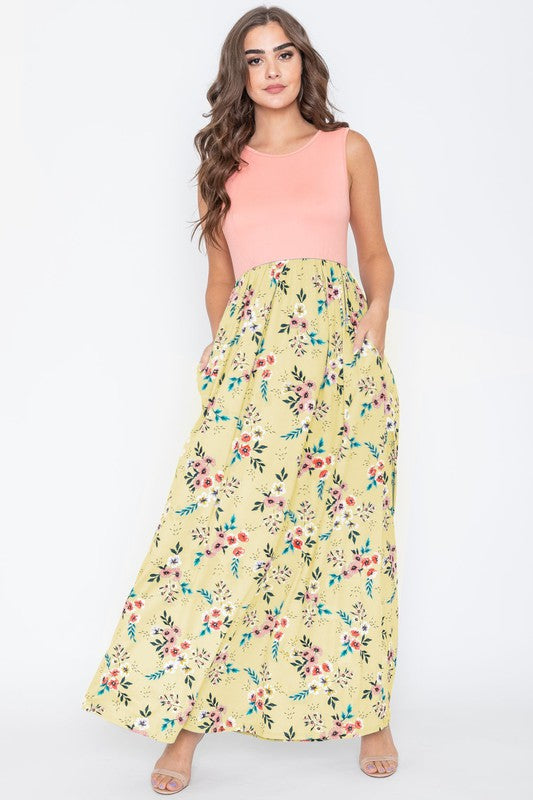 A woman wearing the Summer Floral Maxi Dress, which features a sleeveless design with a pink top and a yellow floral skirt, stands against a plain background. This floor-length dress is proudly made in the United States.