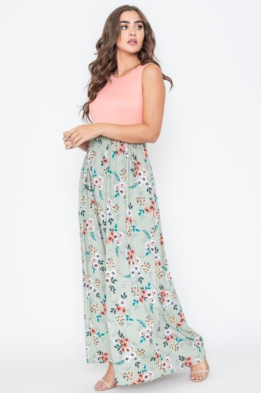 A woman wearing the Summer Floral Maxi Dress, which features a sleeveless design with a pink top and a yellow floral skirt, stands against a plain background. This floor-length dress is proudly made in the United States.