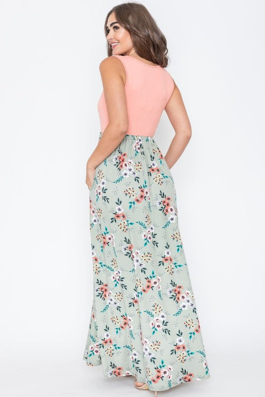 A woman wearing the Summer Floral Maxi Dress, which features a sleeveless design with a pink top and a yellow floral skirt, stands against a plain background. This floor-length dress is proudly made in the United States.