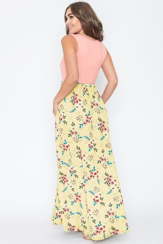 A woman wearing the Summer Floral Maxi Dress, which features a sleeveless design with a pink top and a yellow floral skirt, stands against a plain background. This floor-length dress is proudly made in the United States.