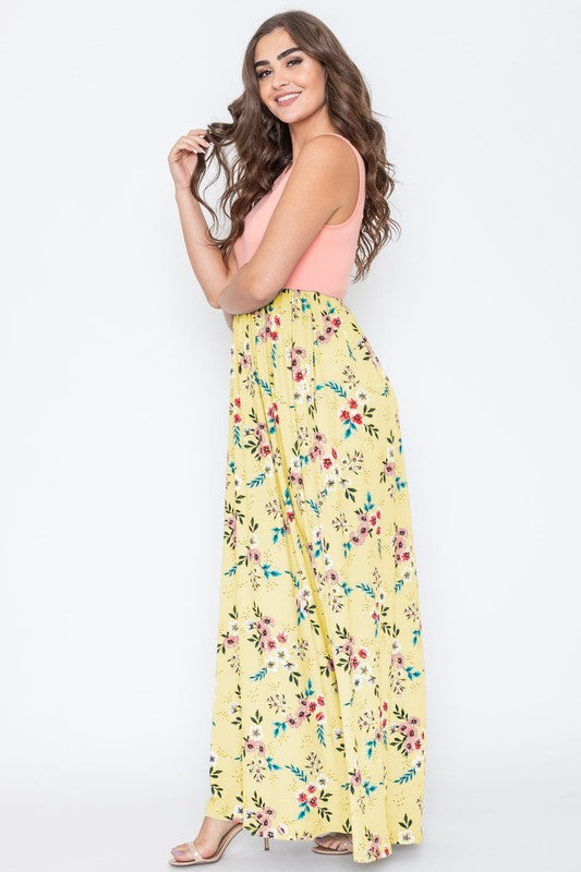 A woman wearing the Summer Floral Maxi Dress, which features a sleeveless design with a pink top and a yellow floral skirt, stands against a plain background. This floor-length dress is proudly made in the United States.