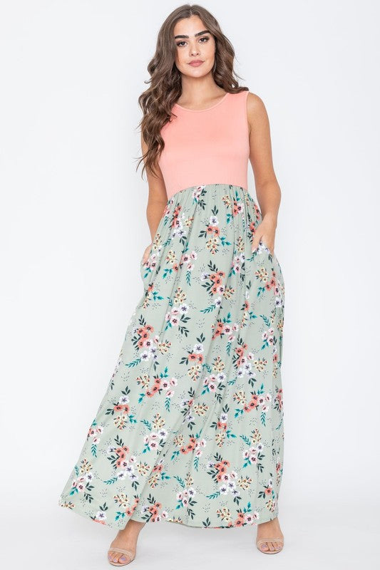 A woman wearing the Summer Floral Maxi Dress, which features a sleeveless design with a pink top and a yellow floral skirt, stands against a plain background. This floor-length dress is proudly made in the United States.