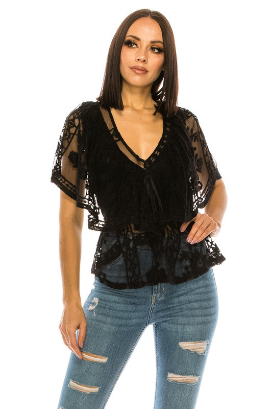 A woman with straight dark hair is wearing the Lace Black Top, characterized by its sheer fabric, V-neck design, and floral pattern. She pairs it with ripped blue jeans as she stands and looks slightly to the side against a plain white background.