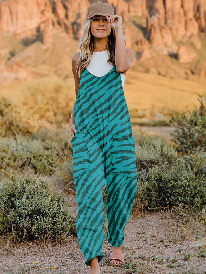 A person wearing a casual white V-neck tank top and a Full Size Printed V-Neck Sleeveless Jumpsuit with a brown animal print zebra-stripe pattern stands outdoors with one hand on their hat and the other in their pocket, set against a rocky landscape backdrop.