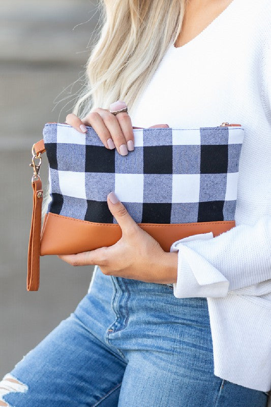 A person in a white sweater and ripped jeans holds the Buffalo Plaid Clutch, a checkered and tan leather accessory with a handle, featuring a stylish buffalo plaid design.