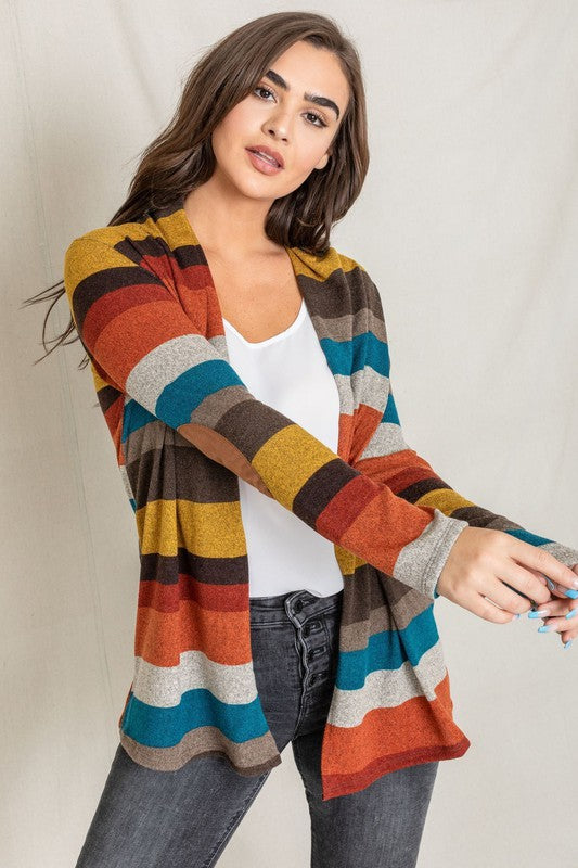 Dressed in a vibrant Stripe Elbow Patch Cardigan composed of soft spandex and polyester, paired with a white top and jeans, an individual stands against a light background.