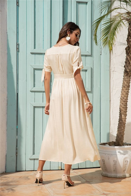 A woman wearing a cream-colored maxi dress with button-front detailing and a smocked waist, paired with sunglasses, stands against a white background.