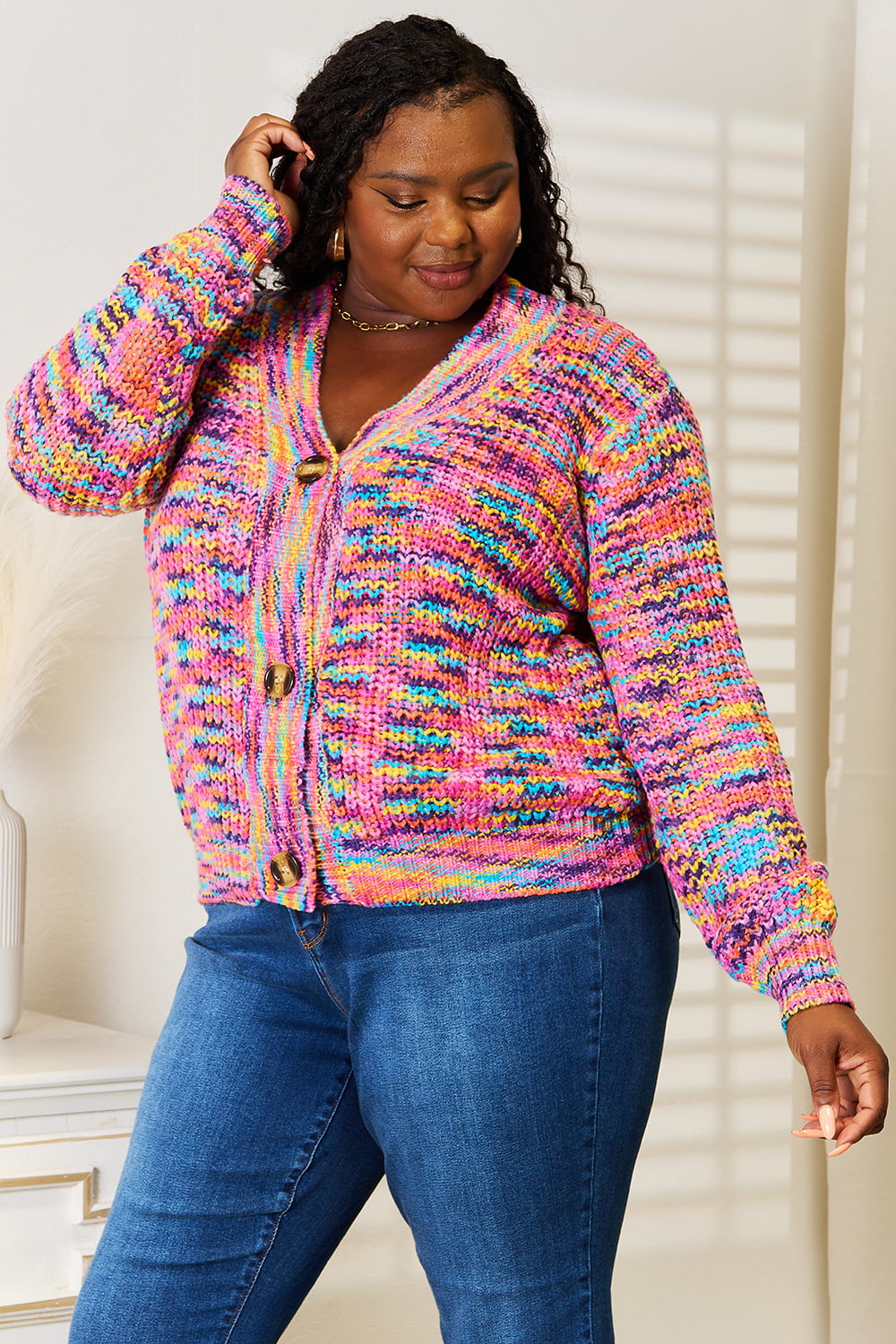 Person wearing an Angel Wings Woven Right V-Neck Long Sleeve Cardigan, characterized by its multicolored design and large buttons, paired with blue jeans. The cardigan, made of 100% acrylic knit, offers a versatile look perfect for any occasion. They are standing indoors against a white background.