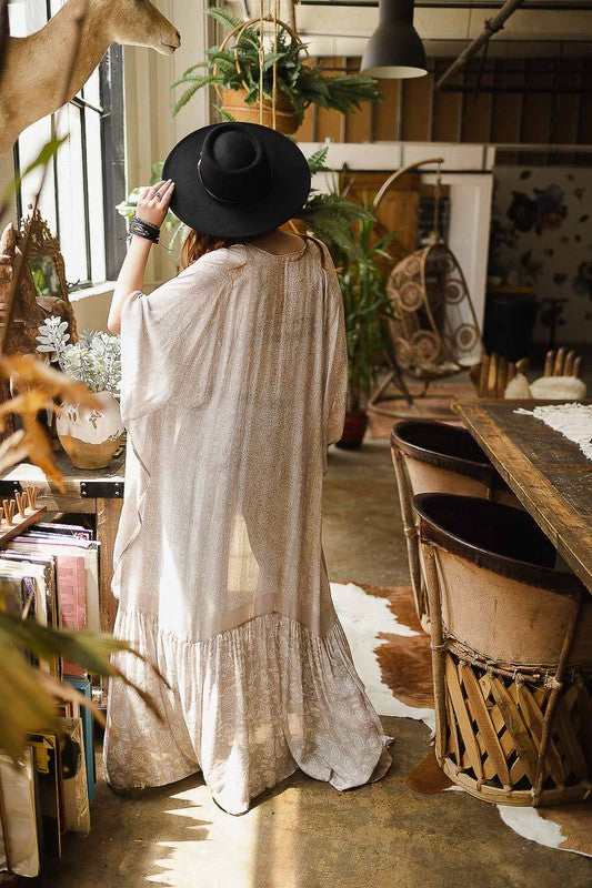 A woman in a long, light-colored Paisley Tapestry Free Flow Kimono and wide-brimmed hat stands in a sunlit room filled with eclectic decor, plants, and vintage items. A deer head is mounted on the wall above her.