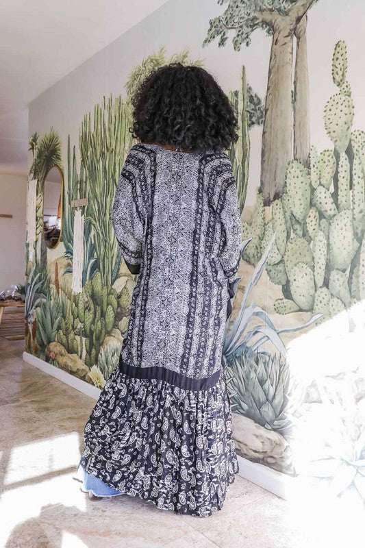 A woman wearing a Paisley Tapestry Free Flow Kimono stands in a bright, eclectic room with plants, books, and an oversized deer head on the wall. The space features large windows and industrial decor.