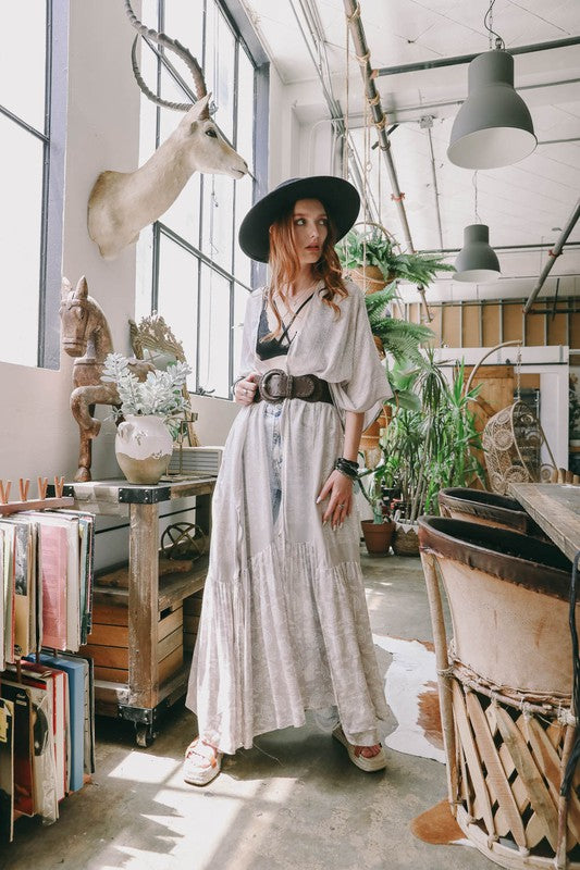 A woman wearing a Paisley Tapestry Free Flow Kimono stands in a bright, eclectic room with plants, books, and an oversized deer head on the wall. The space features large windows and industrial decor.