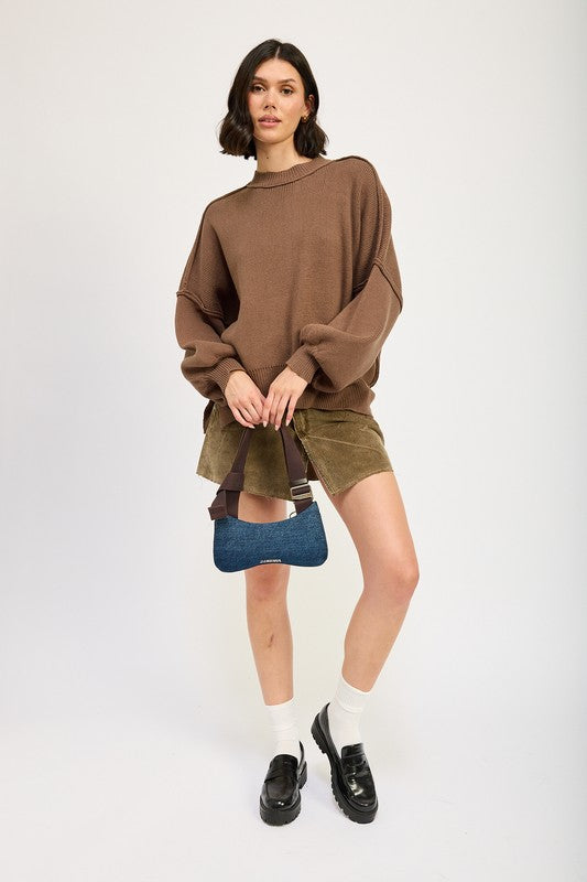 A woman wearing an OVER FITTED LONG SLEEVE SWEATER TOP made of 60% cotton in brown and khaki shorts holds a denim handbag with a brown strap against a neutral background.