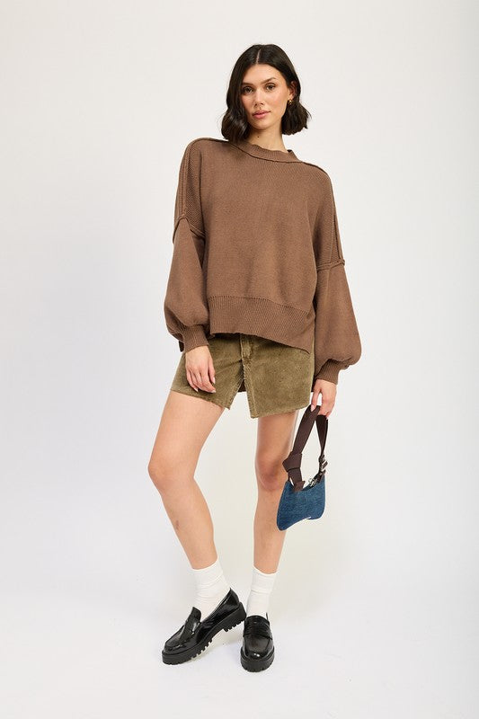 A woman wearing an OVER FITTED LONG SLEEVE SWEATER TOP made of 60% cotton in brown and khaki shorts holds a denim handbag with a brown strap against a neutral background.