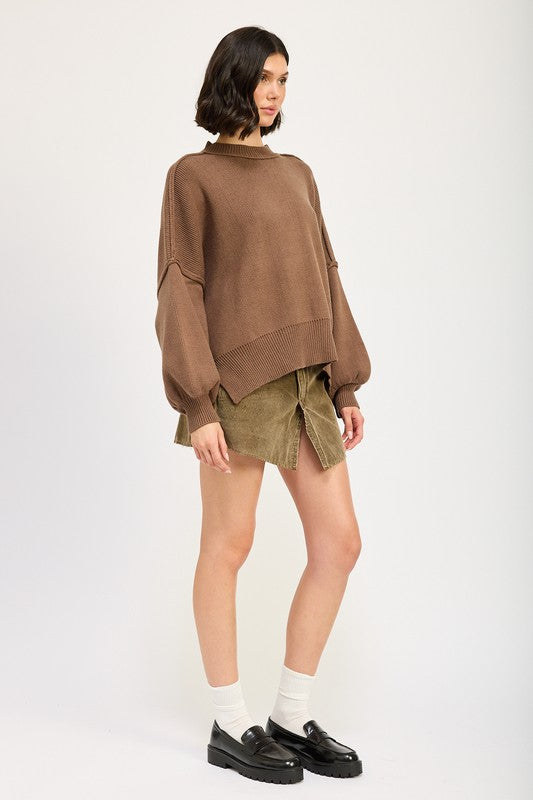 A woman wearing an OVER FITTED LONG SLEEVE SWEATER TOP made of 60% cotton in brown and khaki shorts holds a denim handbag with a brown strap against a neutral background.