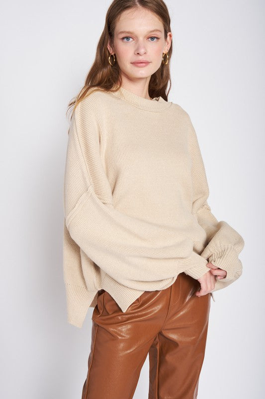 A woman wearing an OVER FITTED LONG SLEEVE SWEATER TOP made of 60% cotton in brown and khaki shorts holds a denim handbag with a brown strap against a neutral background.