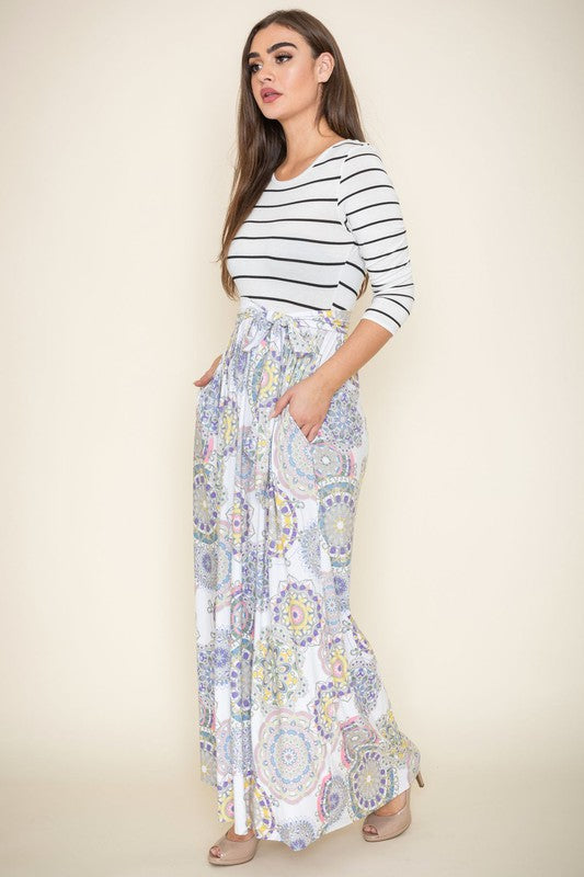 A woman wearing a Sash Maxi Dress With Pockets, reminiscent of a striped top and floral-patterned skirt design, stands against a beige background.