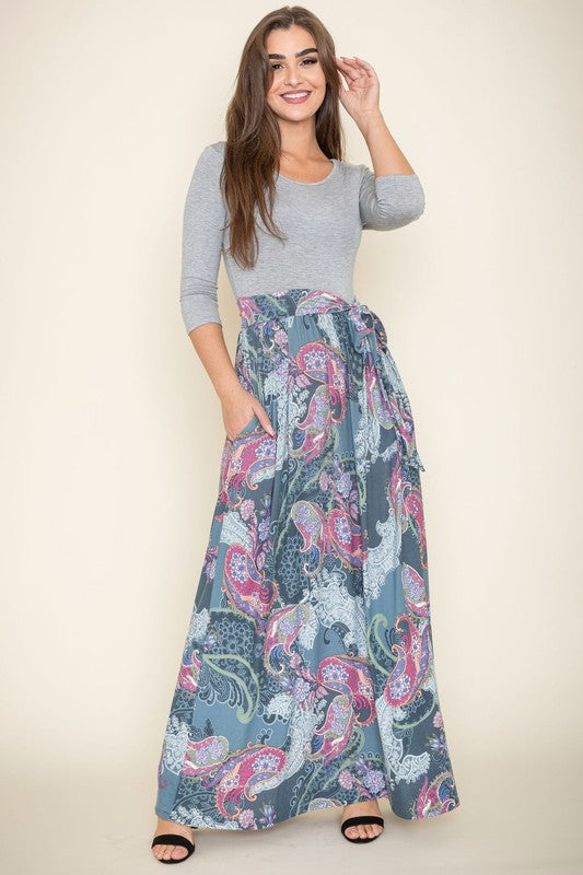 A woman in a Plus Sash Maxi Dress with Pockets, featuring long sleeves in gray and a long, colorful paisley-patterned skirt, stands against a beige backdrop.