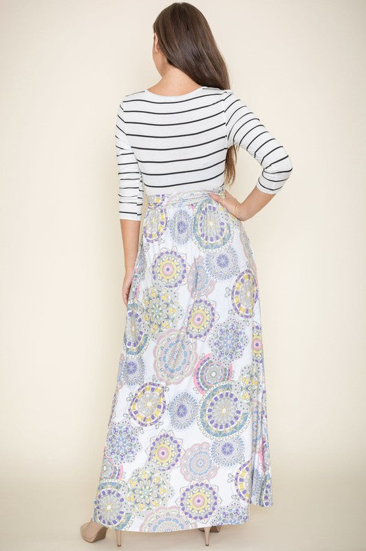 A woman in a Plus Sash Maxi Dress with Pockets, featuring long sleeves in gray and a long, colorful paisley-patterned skirt, stands against a beige backdrop.