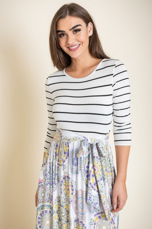 A woman wearing a Sash Maxi Dress With Pockets, reminiscent of a striped top and floral-patterned skirt design, stands against a beige background.