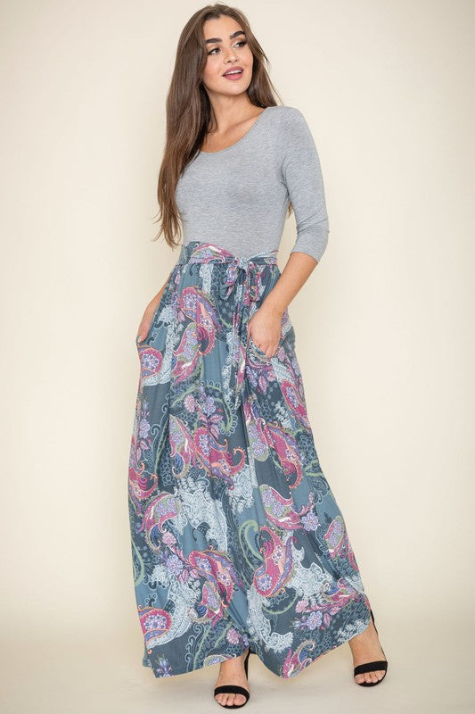 A woman wearing a Sash Maxi Dress With Pockets, reminiscent of a striped top and floral-patterned skirt design, stands against a beige background.