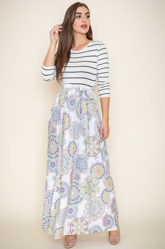 A woman in a Plus Sash Maxi Dress with Pockets, featuring long sleeves in gray and a long, colorful paisley-patterned skirt, stands against a beige backdrop.