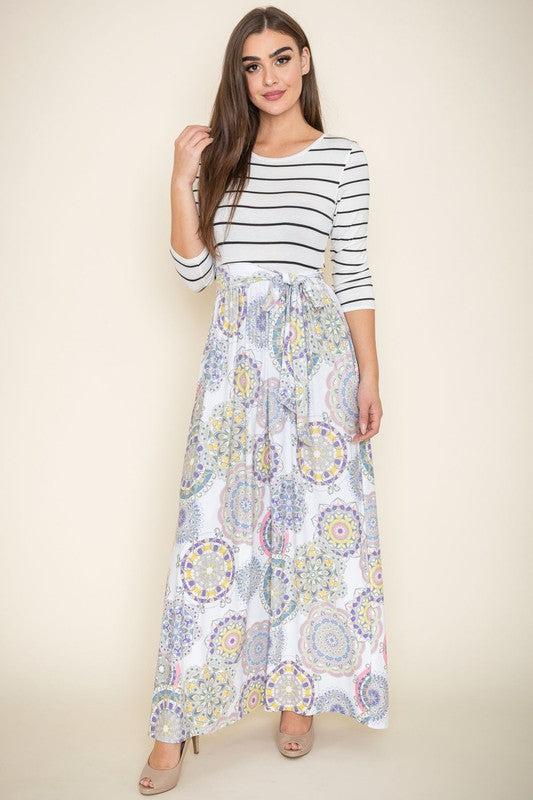 A woman wearing a Sash Maxi Dress With Pockets, reminiscent of a striped top and floral-patterned skirt design, stands against a beige background.