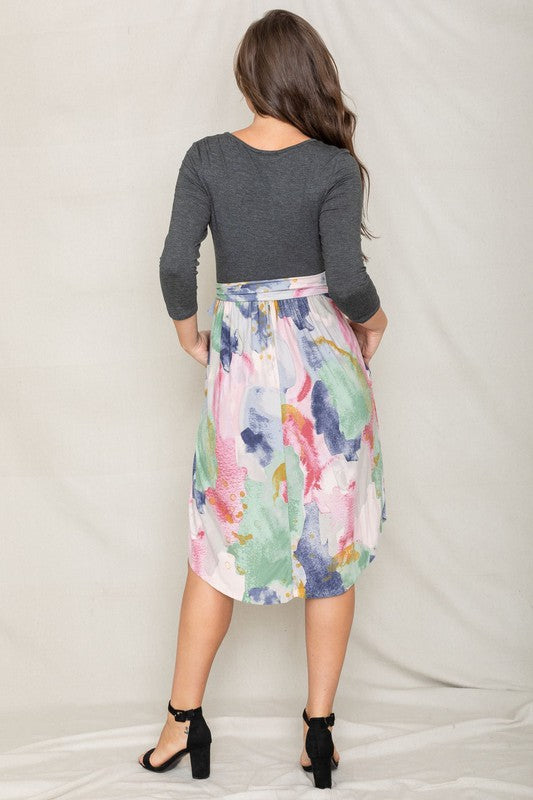 A woman in a gray long-sleeve top and the Watercolor Sash Midi Dress stands elegantly against a plain background.