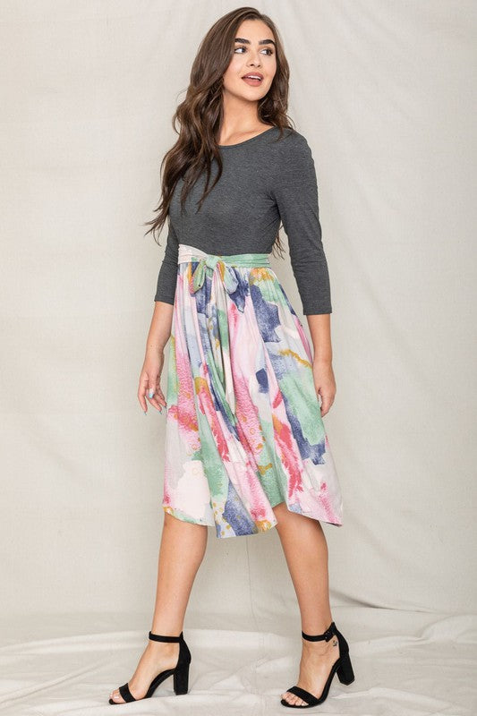 A woman in a gray long-sleeve top and the Watercolor Sash Midi Dress stands elegantly against a plain background.