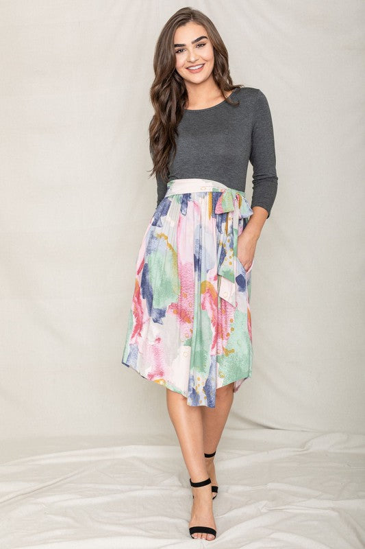 A woman in a gray long-sleeve top and the Watercolor Sash Midi Dress stands elegantly against a plain background.