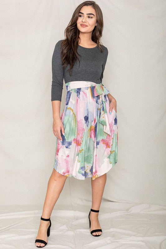 A woman in a gray long-sleeve top and the Watercolor Sash Midi Dress stands elegantly against a plain background.