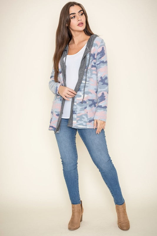 A woman wears a Camo Cardigan with a hoodie over a white top and jeans, standing gracefully against a pale backdrop, effortlessly blending casual style with modern flair.