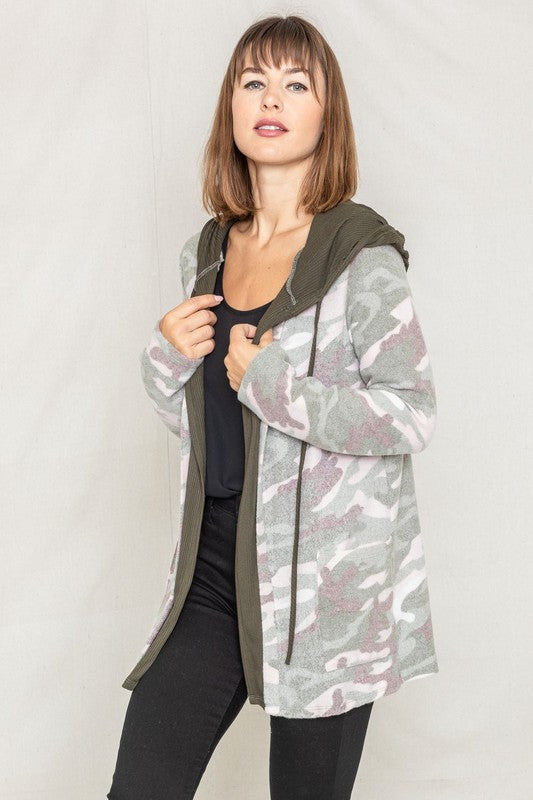 A woman wears a Camo Cardigan with a hoodie over a white top and jeans, standing gracefully against a pale backdrop, effortlessly blending casual style with modern flair.