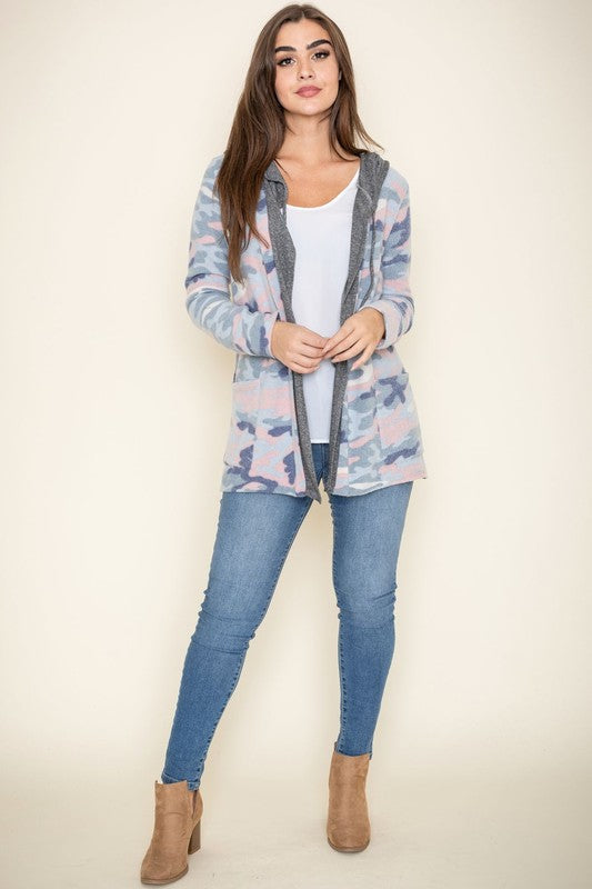 A woman wears a Camo Cardigan with a hoodie over a white top and jeans, standing gracefully against a pale backdrop, effortlessly blending casual style with modern flair.