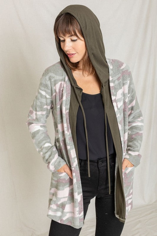 A woman wears a Camo Cardigan with a hoodie over a white top and jeans, standing gracefully against a pale backdrop, effortlessly blending casual style with modern flair.