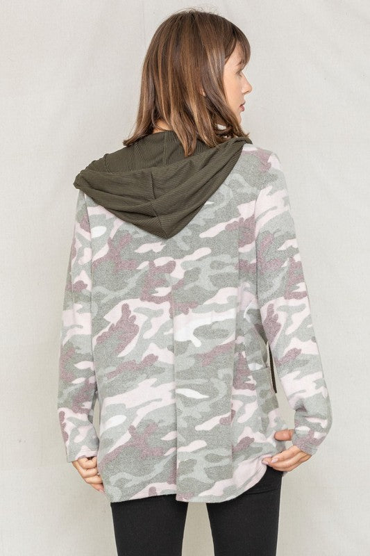 A woman wears a Camo Cardigan with a hoodie over a white top and jeans, standing gracefully against a pale backdrop, effortlessly blending casual style with modern flair.