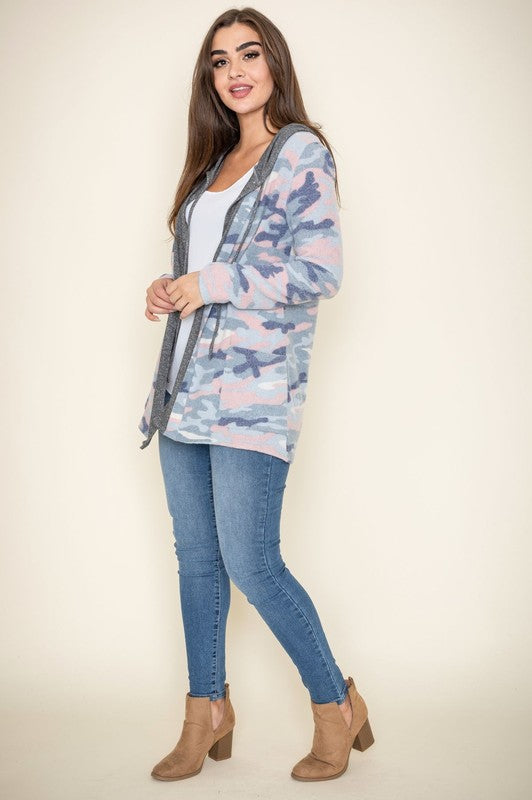 A woman wears a Camo Cardigan with a hoodie over a white top and jeans, standing gracefully against a pale backdrop, effortlessly blending casual style with modern flair.