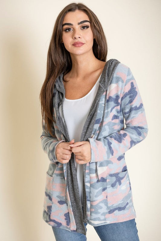 A woman wears a Camo Cardigan with a hoodie over a white top and jeans, standing gracefully against a pale backdrop, effortlessly blending casual style with modern flair.